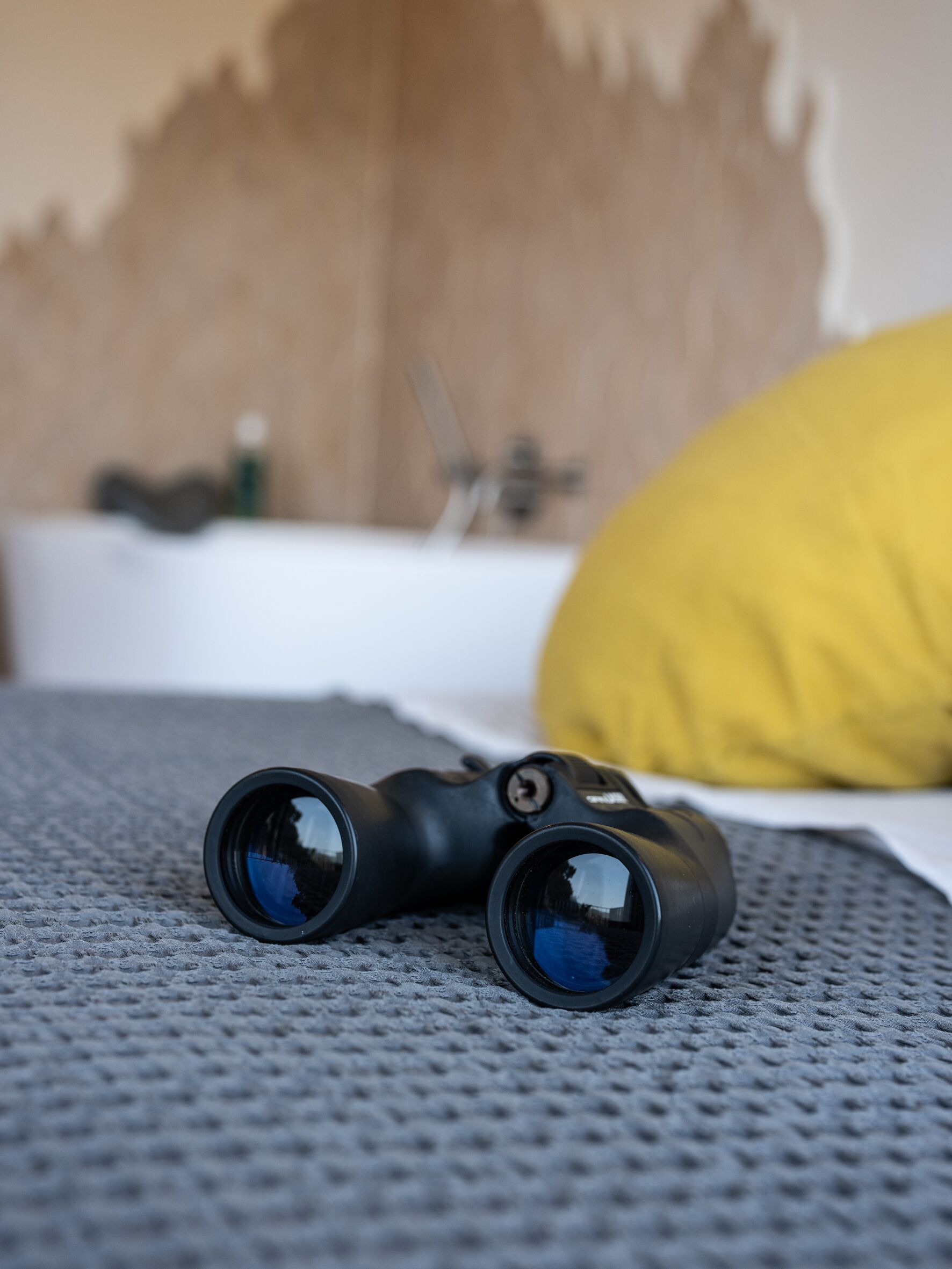 A pair of binoculars on a textured surface, possibly a bed, with a yellow pillow and a wooden headboard in the background.