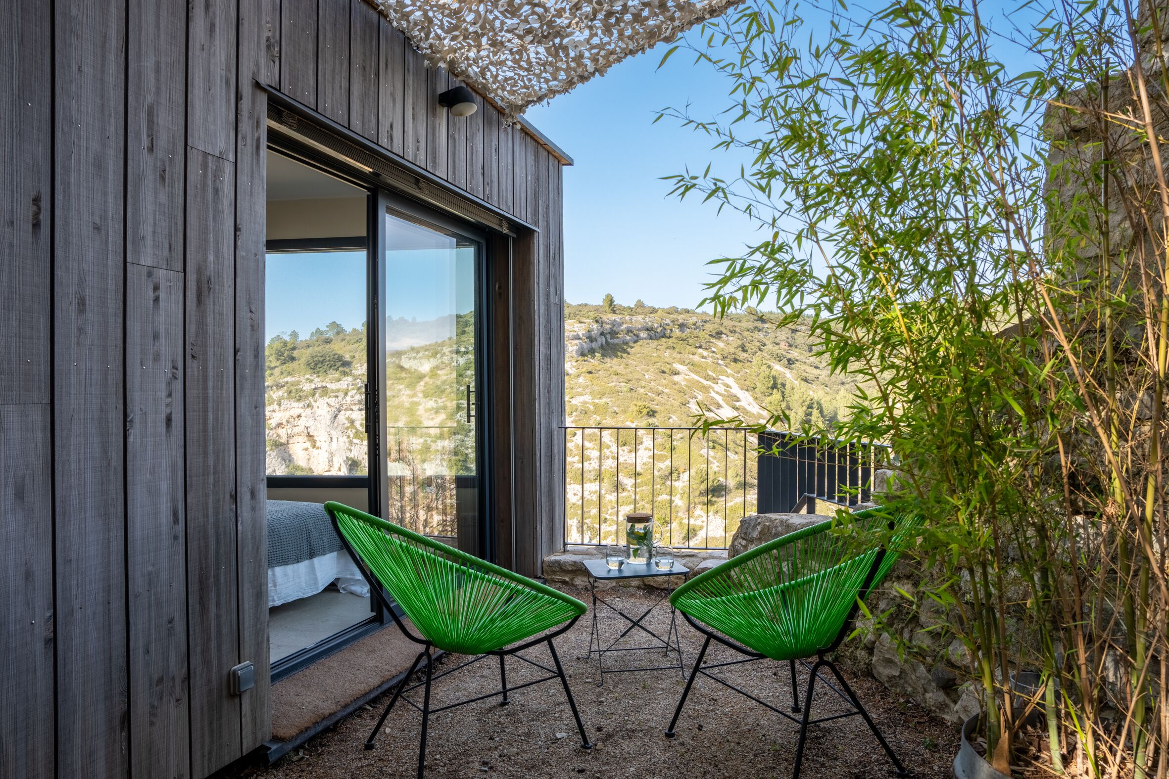 Una construcción de madera con balcón, dos sillas verdes y una mesa con jarrón y vasos.