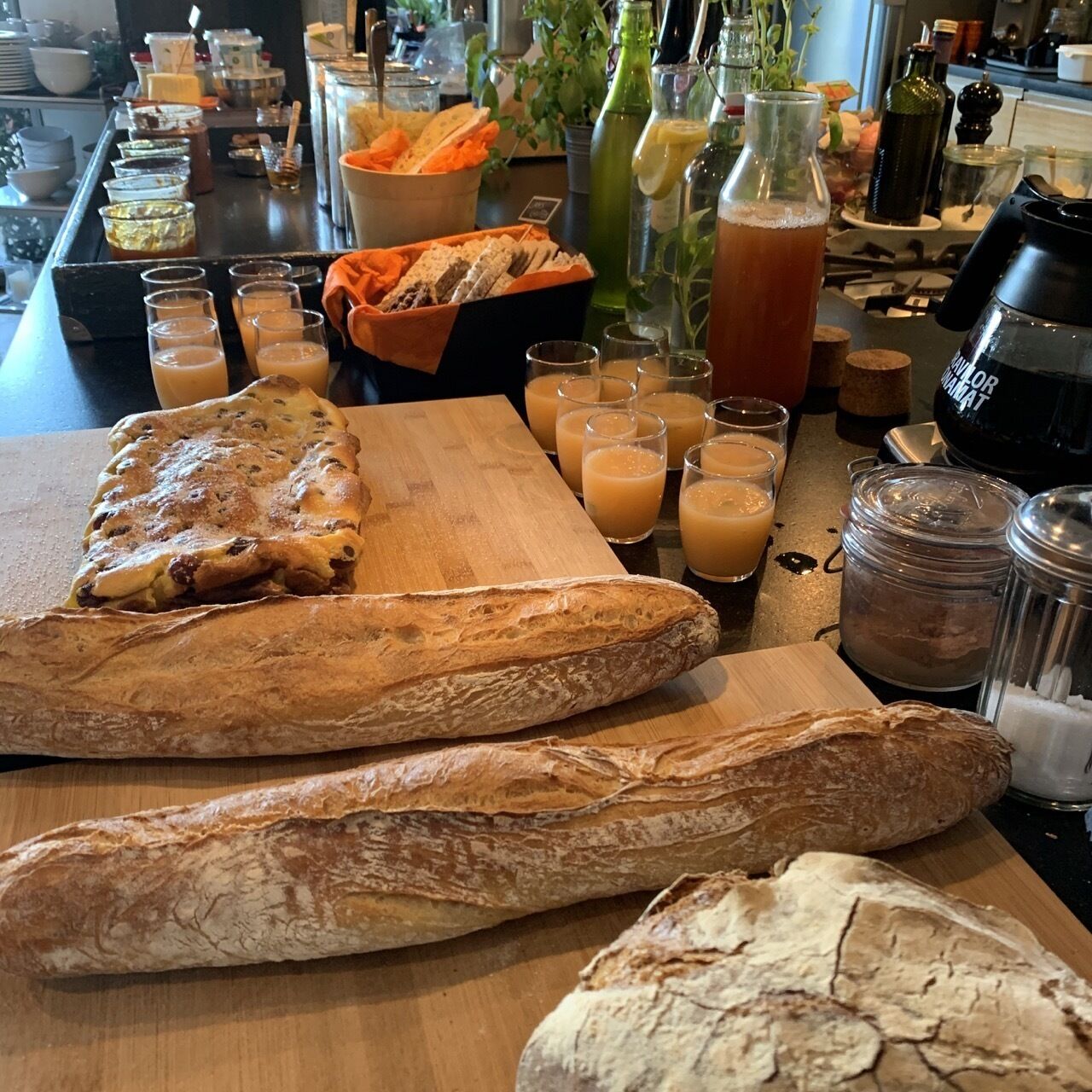 Uma bancada de cozinha com vários pães, potes de condimentos e copos de bebidas.