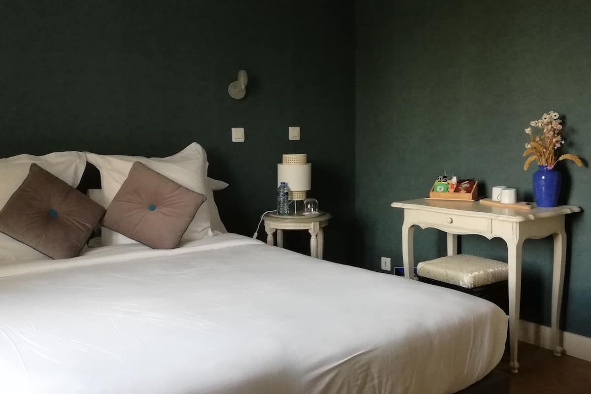 A neatly made bed with white and brown pillows, a bedside table with a lamp, a vase with flowers, and a small desk with a cup and a box.