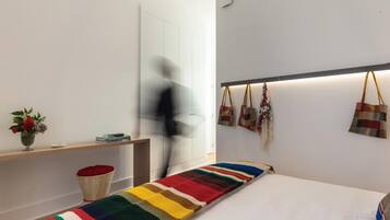 Quarto de hotel moderno com uma cama grande, uma mesa de cabeceira com um vaso de flores, abajur e uma estante na parede com malas.