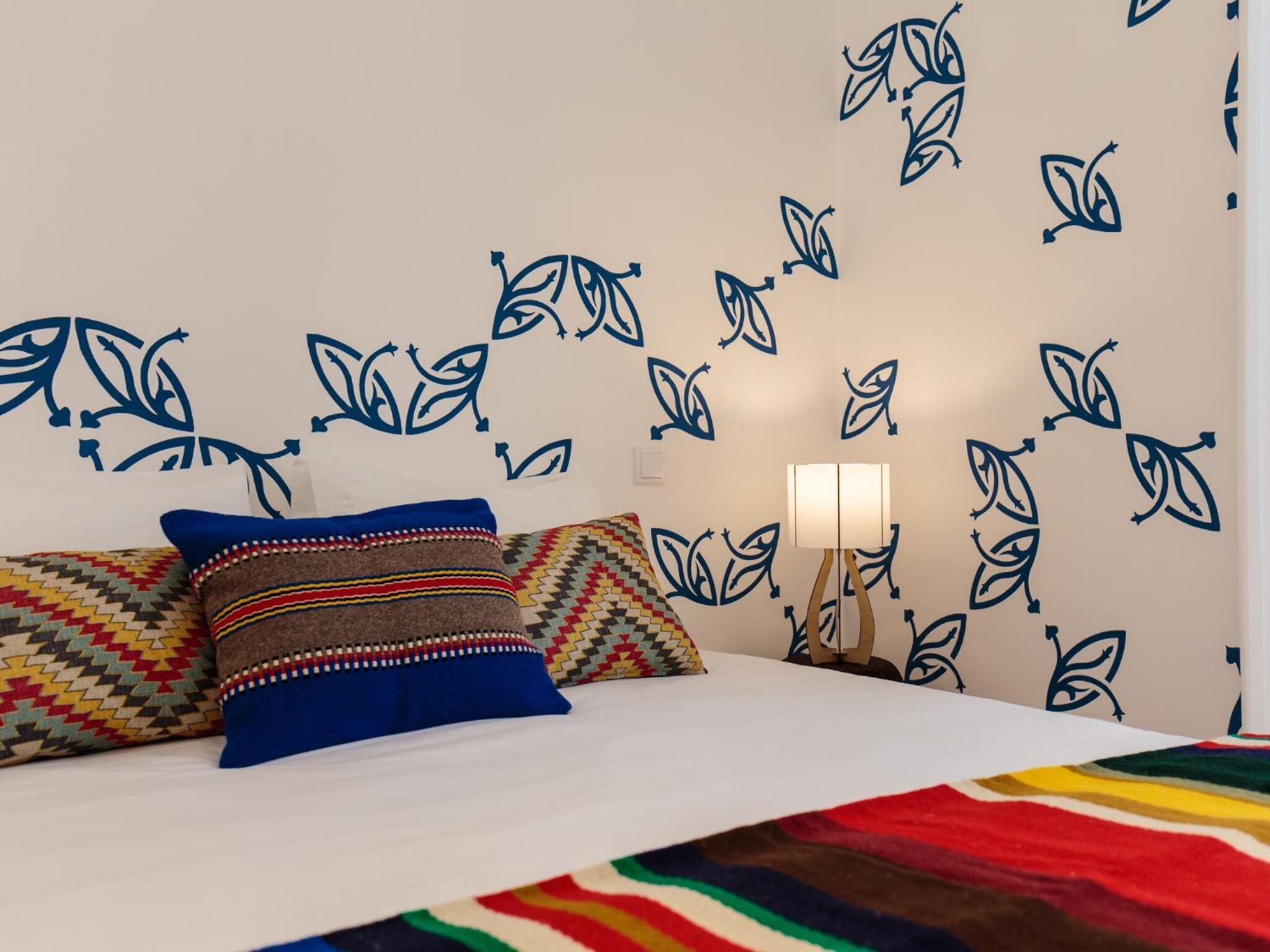 A bed with patterned pillows, a bedside lamp, and a wall with a leaf pattern.
