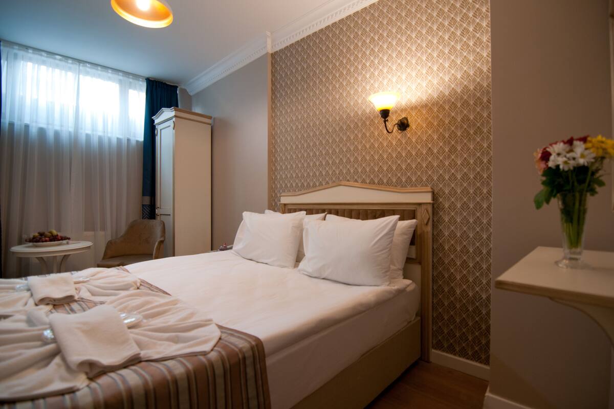 A neatly made bed with white linens, a bedside table with a vase of flowers, and a wall-mounted light fixture.