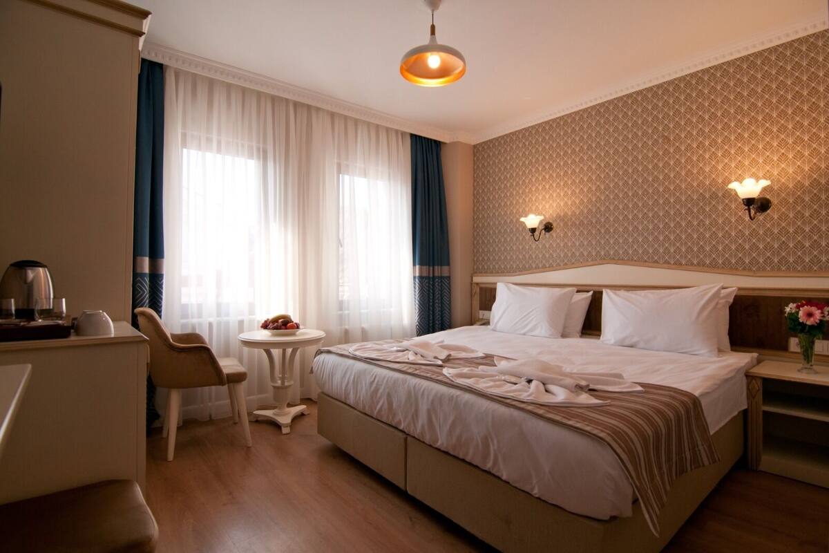 A hotel room with a bed, a desk, a chair, a small table with a fruit bowl, and two wall-mounted lamps.