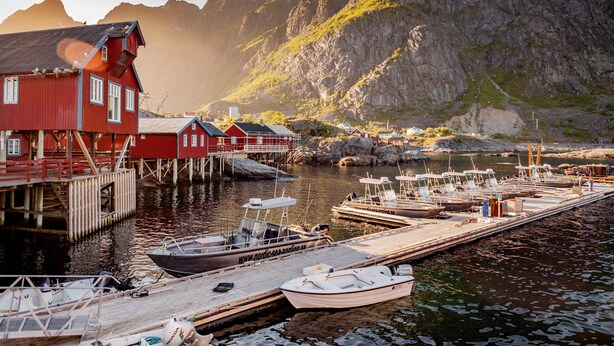 A harbor with boats docked, a red building, and a mountainous backdrop.