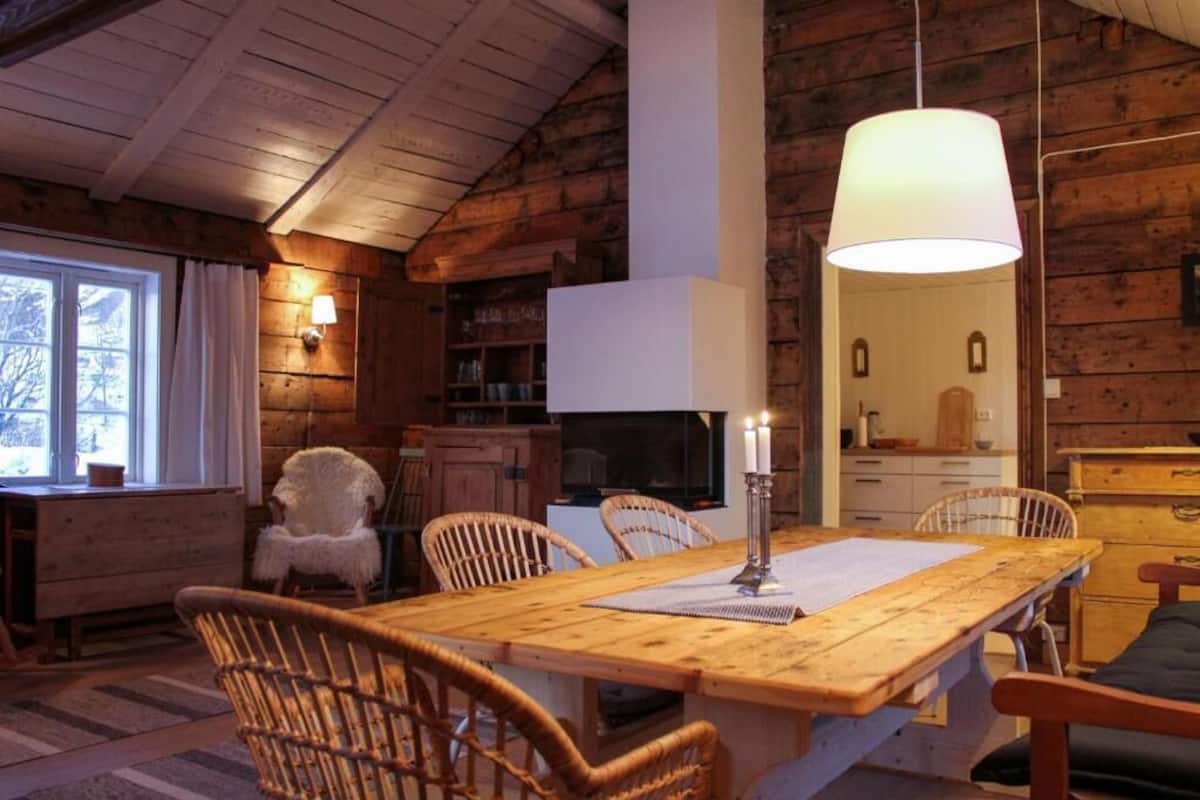 A cozy cabin interior with a wooden dining table, wicker chairs, a fireplace, and a large window.