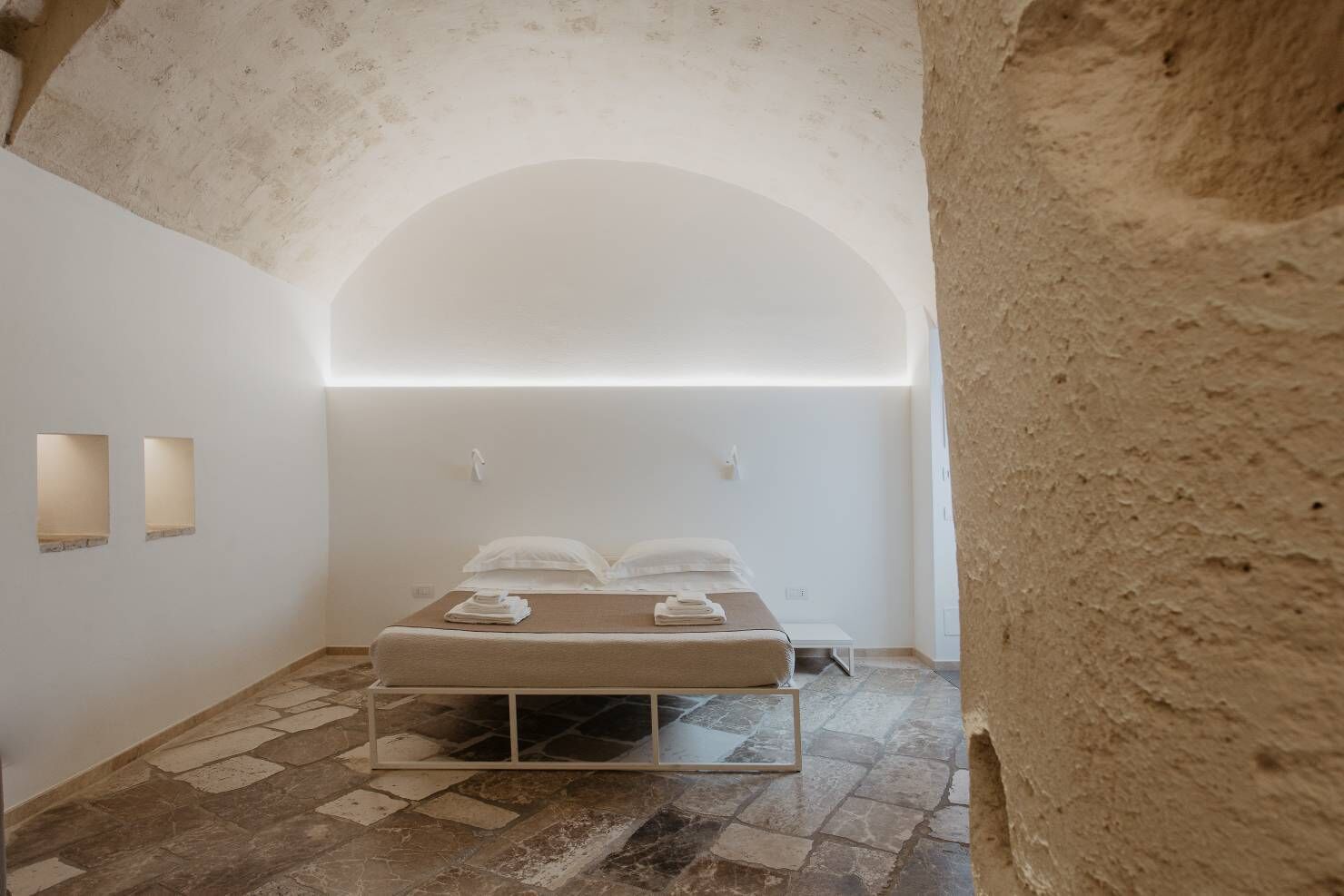 Una habitación estrecha y tenuemente iluminada, con una cama, una mesita y un banco.