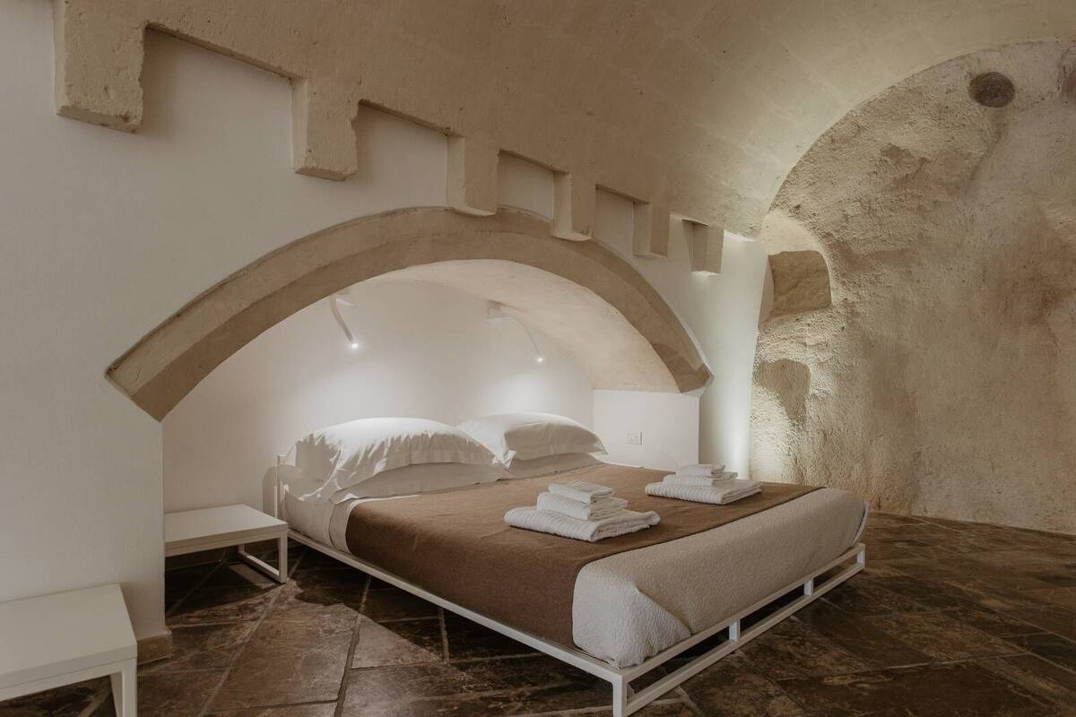 A bedroom with a bed under an arched ceiling, a nightstand, and a large stone wall.