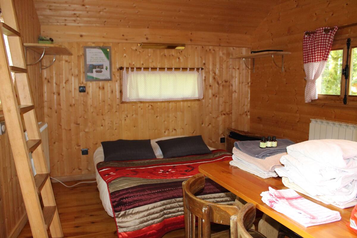 A wooden cabin room with a bed, a table with towels, a chair, a window with curtains, and a shelf with items.