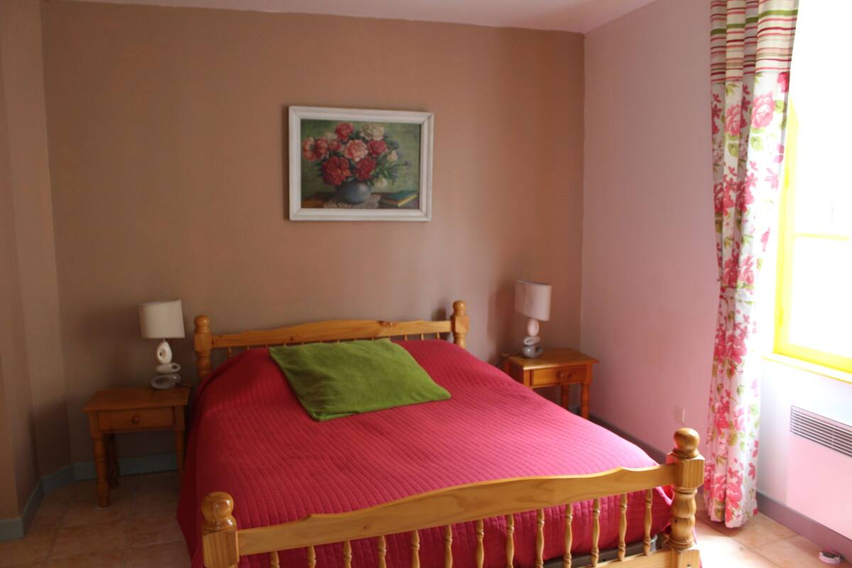 A bedroom with a wooden bed, two bedside tables, a nightstand, a floral painting on the wall, and a window with curtains.