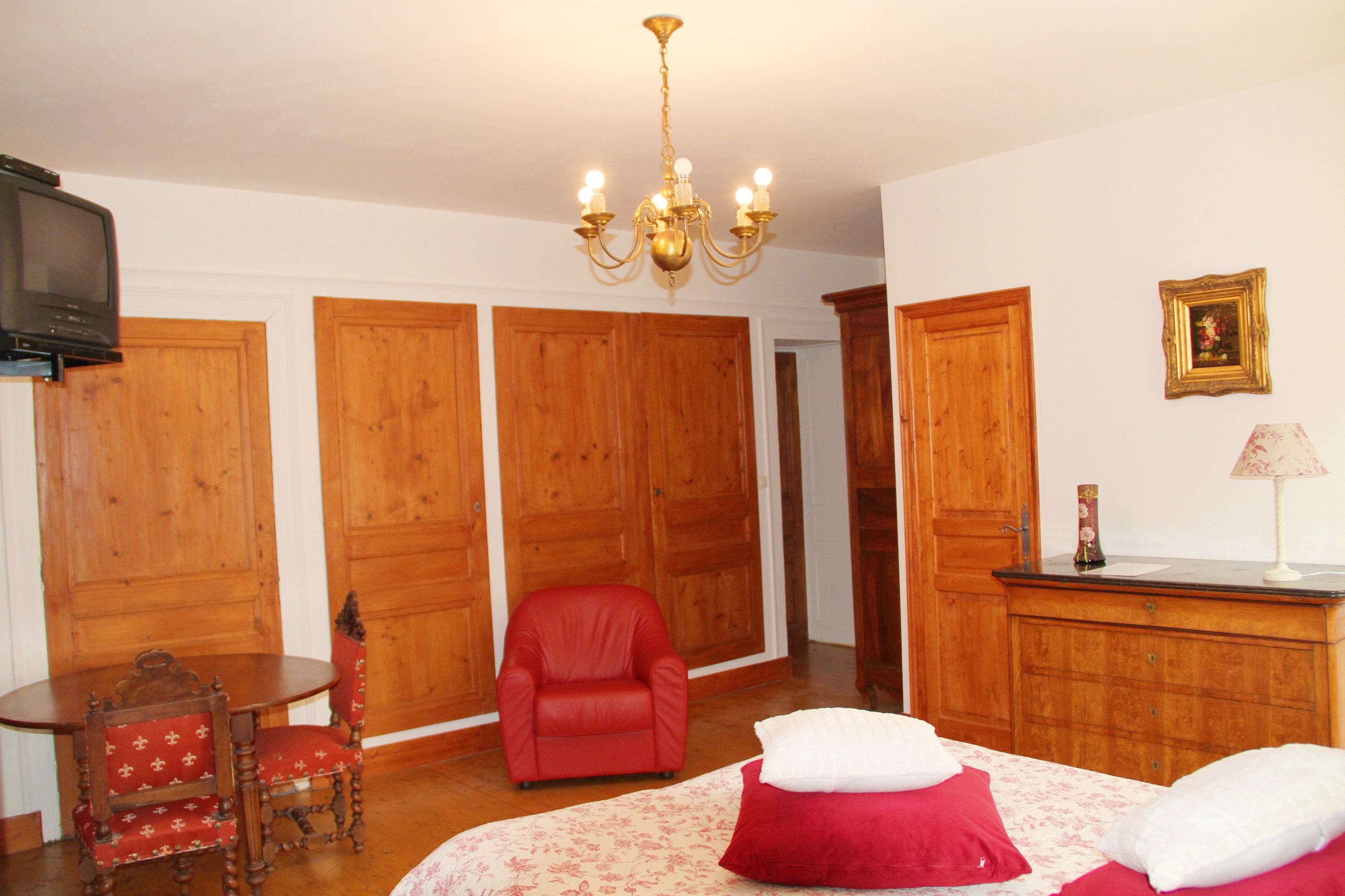 A hotel room with wooden wardrobes, a red armchair, a small round table, and a bed with red and white pillows.