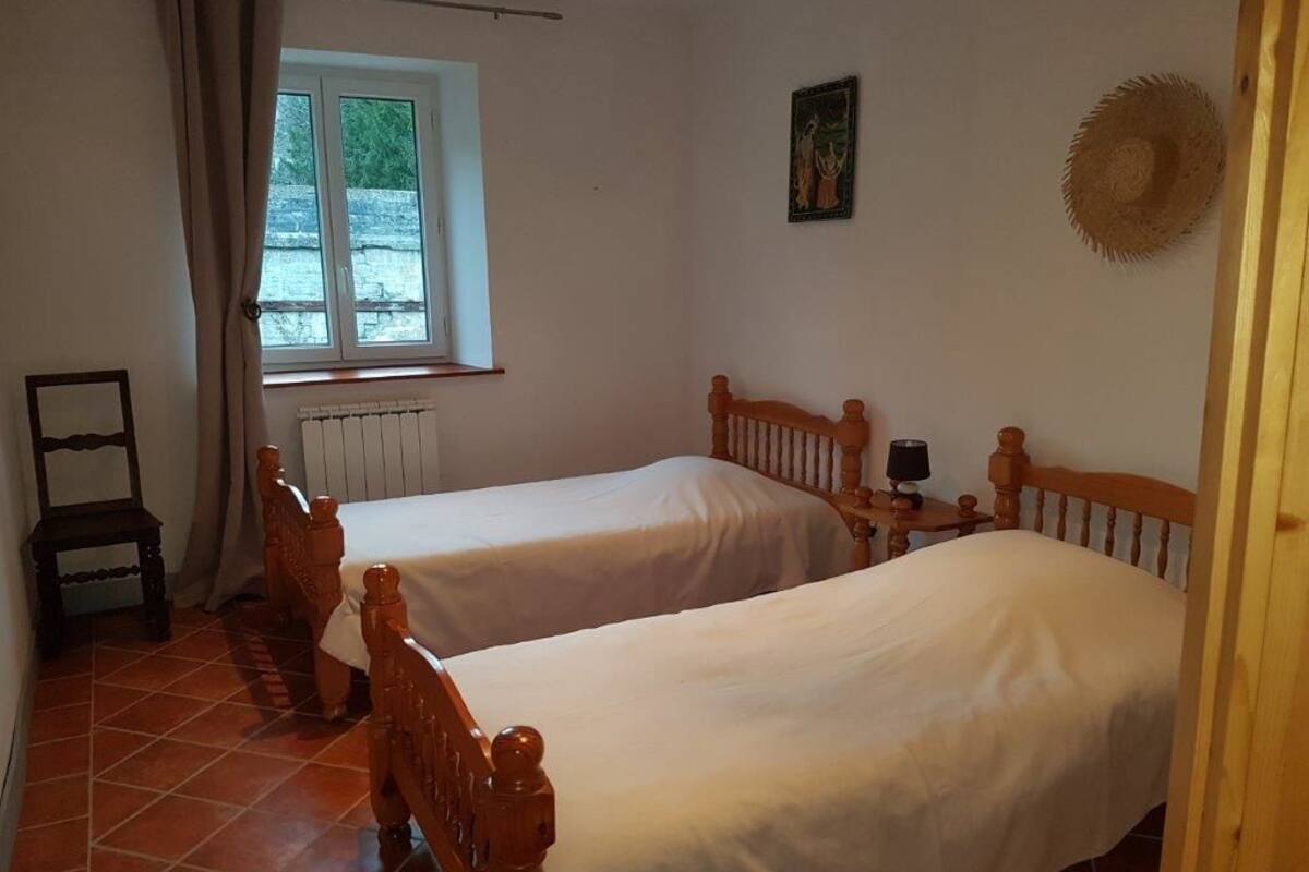 A room with two wooden beds, a radiator, a window with curtains, and a small table with a lamp.