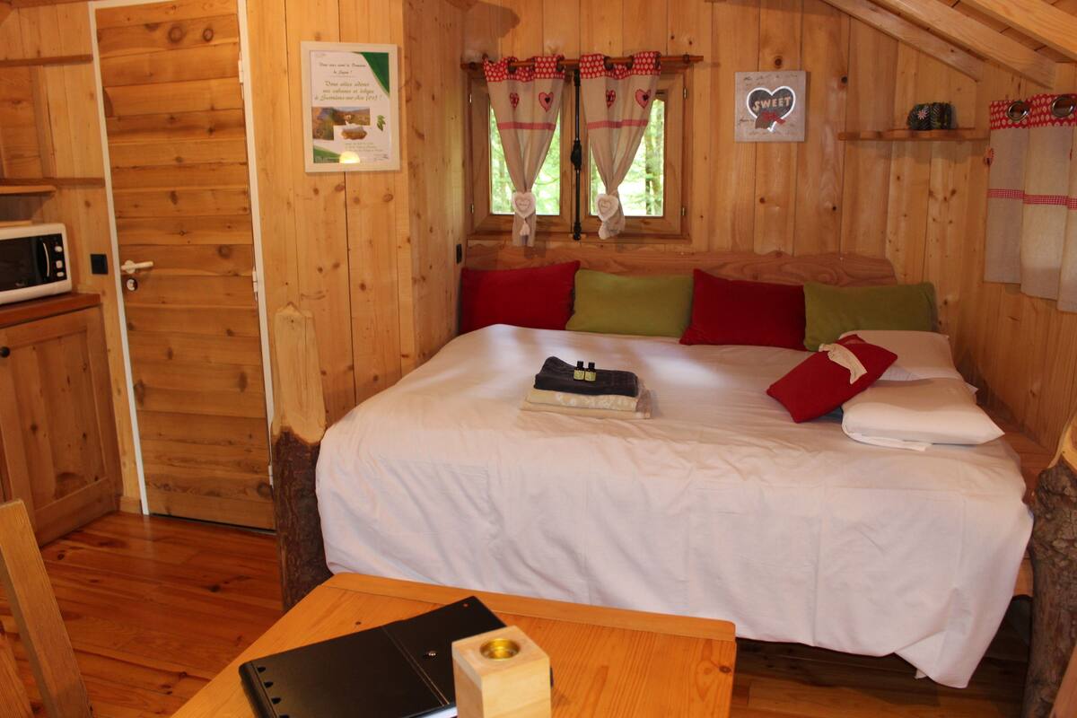 A wooden cabin bedroom with a bed, pillows, a table with a laptop, and a microwave.