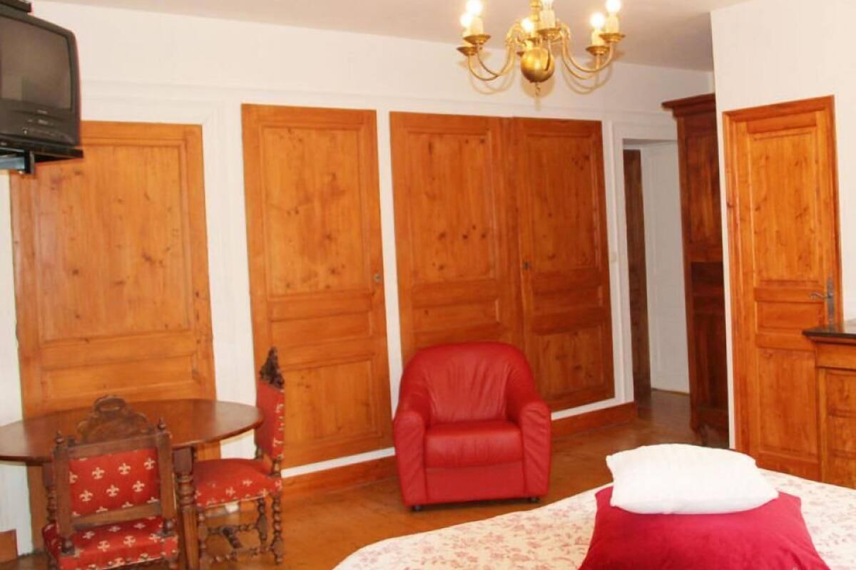 A room with wooden wardrobes, a red armchair, a wooden table with a chair, and a bed with a red pillow.