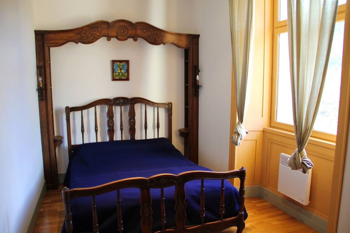 A bedroom with a wooden bed frame, a blue bedspread, a small painting on the wall, and a window with curtains.