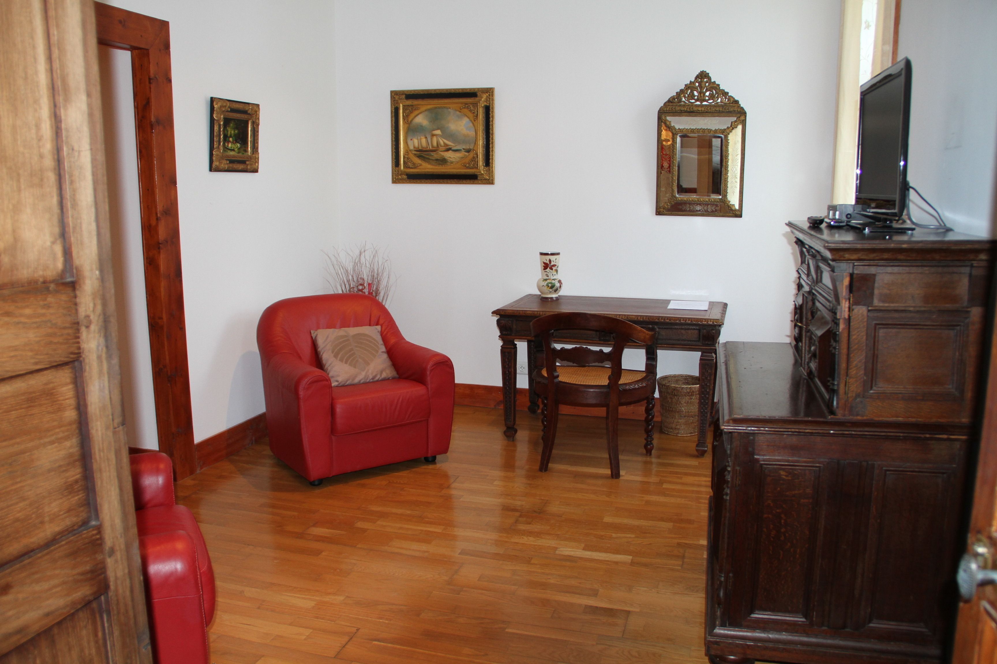 Une pièce avec un plancher en bois, un fauteuil rouge, une table en bois avec une chaise, un meuble en bois et un téléviseur posé sur le dessus.