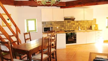 Un coin repas avec une table et des chaises en bois, une cuisine avec des meubles blancs et un dosseret carrelé, et un escalier avec des poutres en bois.