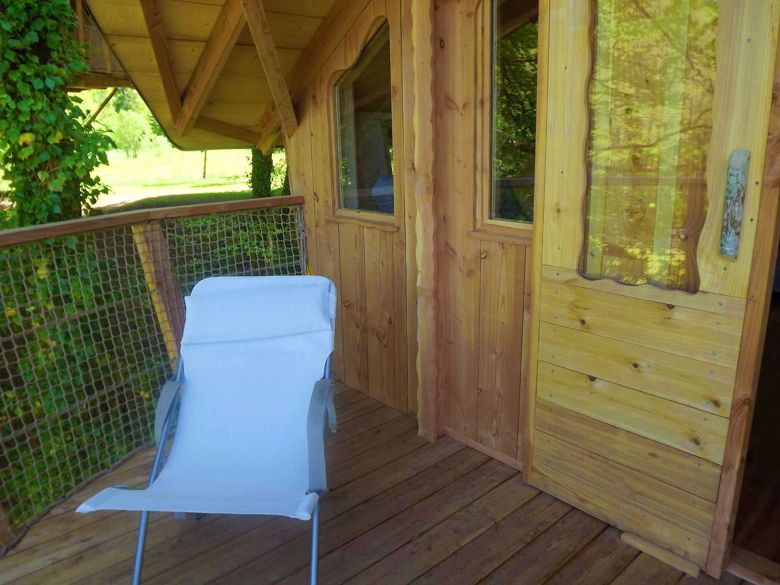 Una cabaña de madera con un porche cubierto, una silla de descanso blanca y una ventana con vistas a la vegetación.