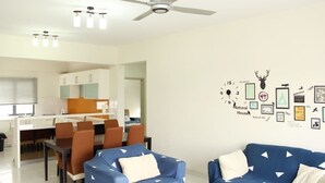 A modern living room with a ceiling fan, a dining area with a white table and brown chairs, and a wall decorated with various framed pictures and a clock. - Setia Walk by SYNC (Puchong)