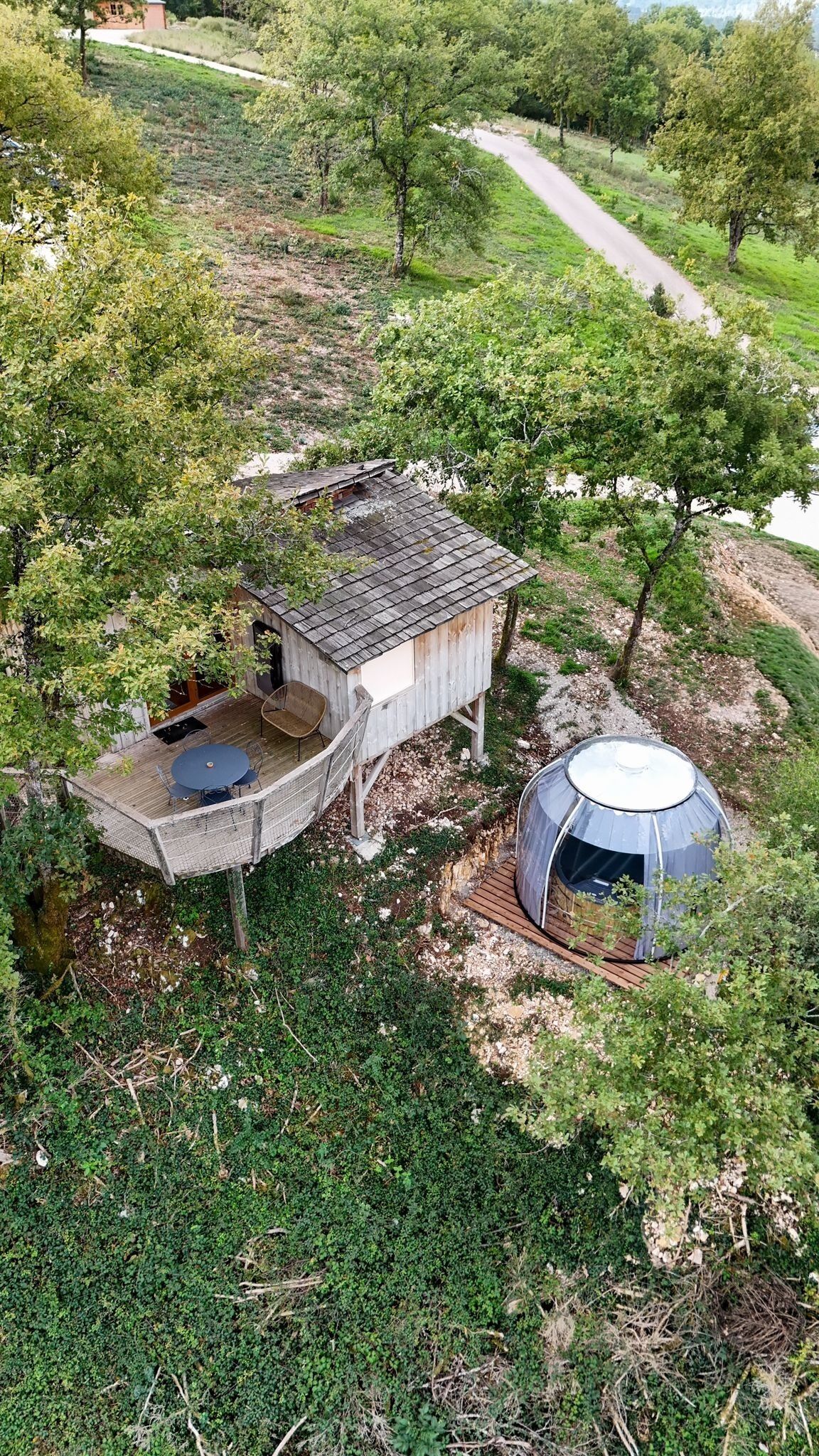 Una cabaña rústica con una pequeña terraza y una estructura en forma de cúpula, rodeada de árboles y un sendero de tierra.