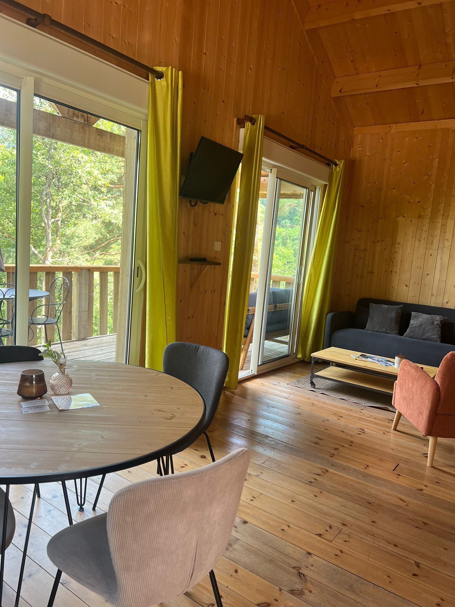 A wooden interior with a round table, chairs, a sofa, and a TV mounted on the wall.