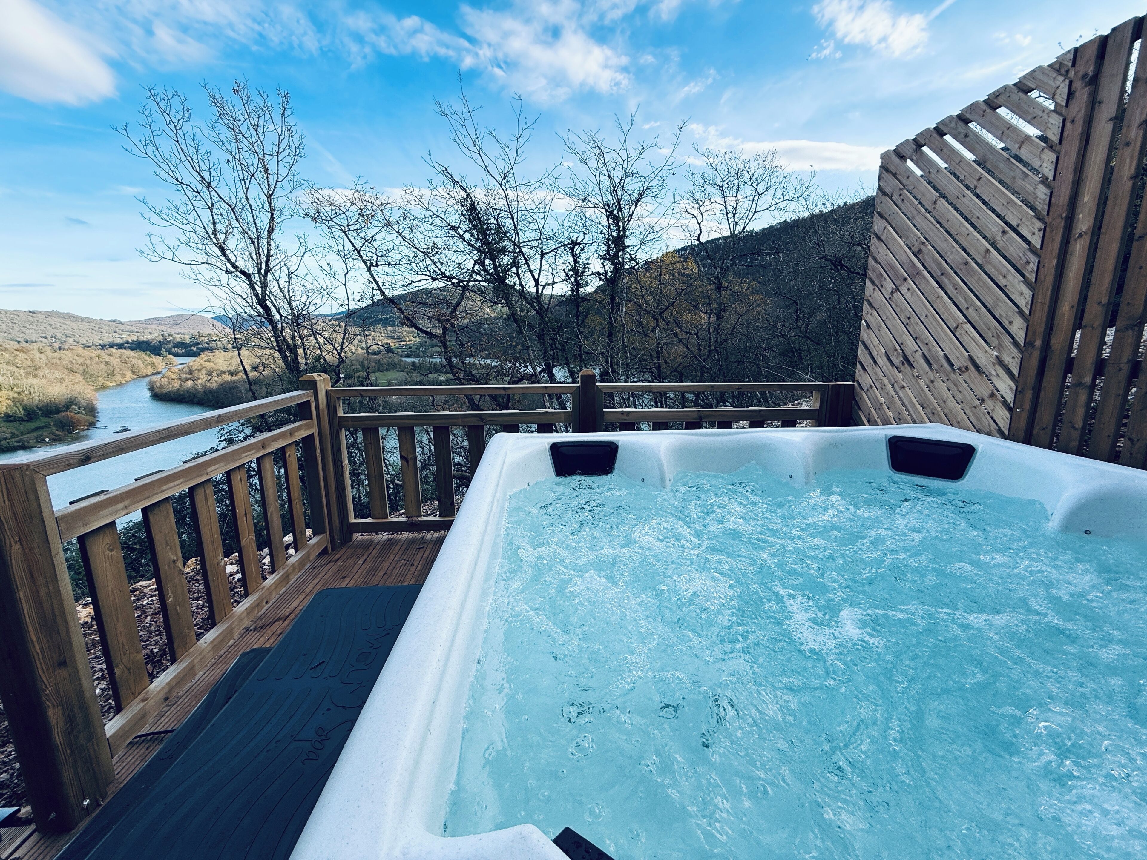 A hot tub on a wooden deck with a river and hills in the background.