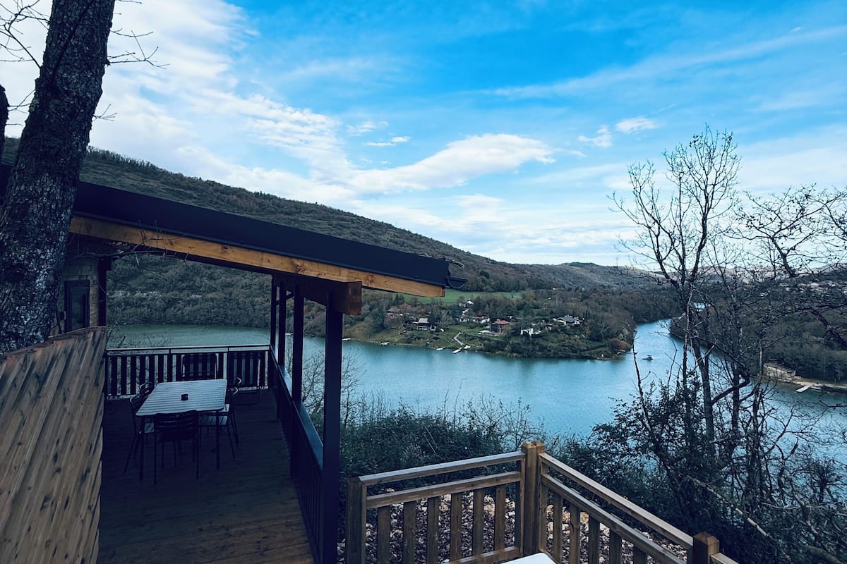 Une terrasse en bois avec un jacuzzi donnant sur un lac et des collines.