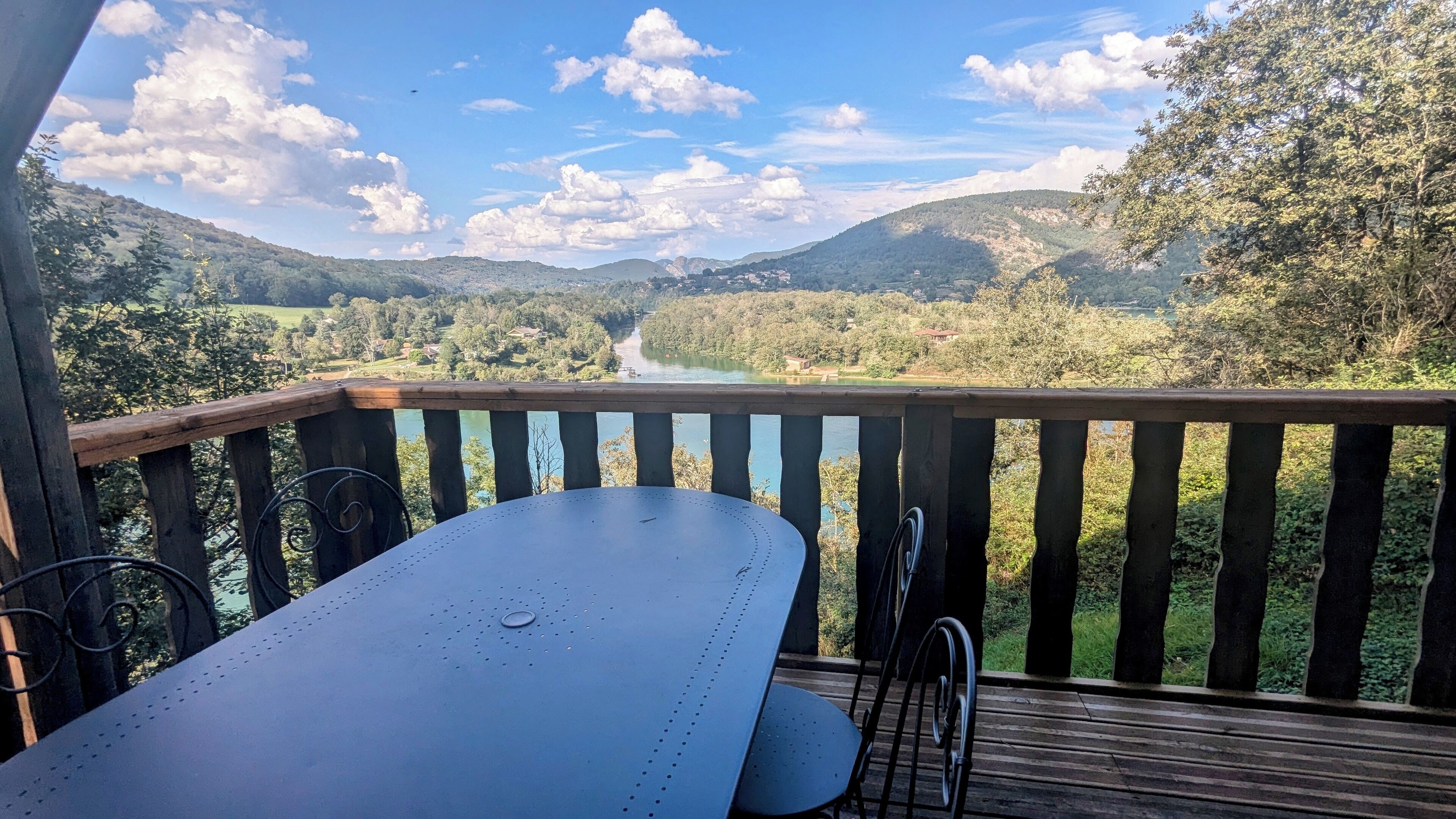 A balcony with a table and chairs overlooking a river and mountains.