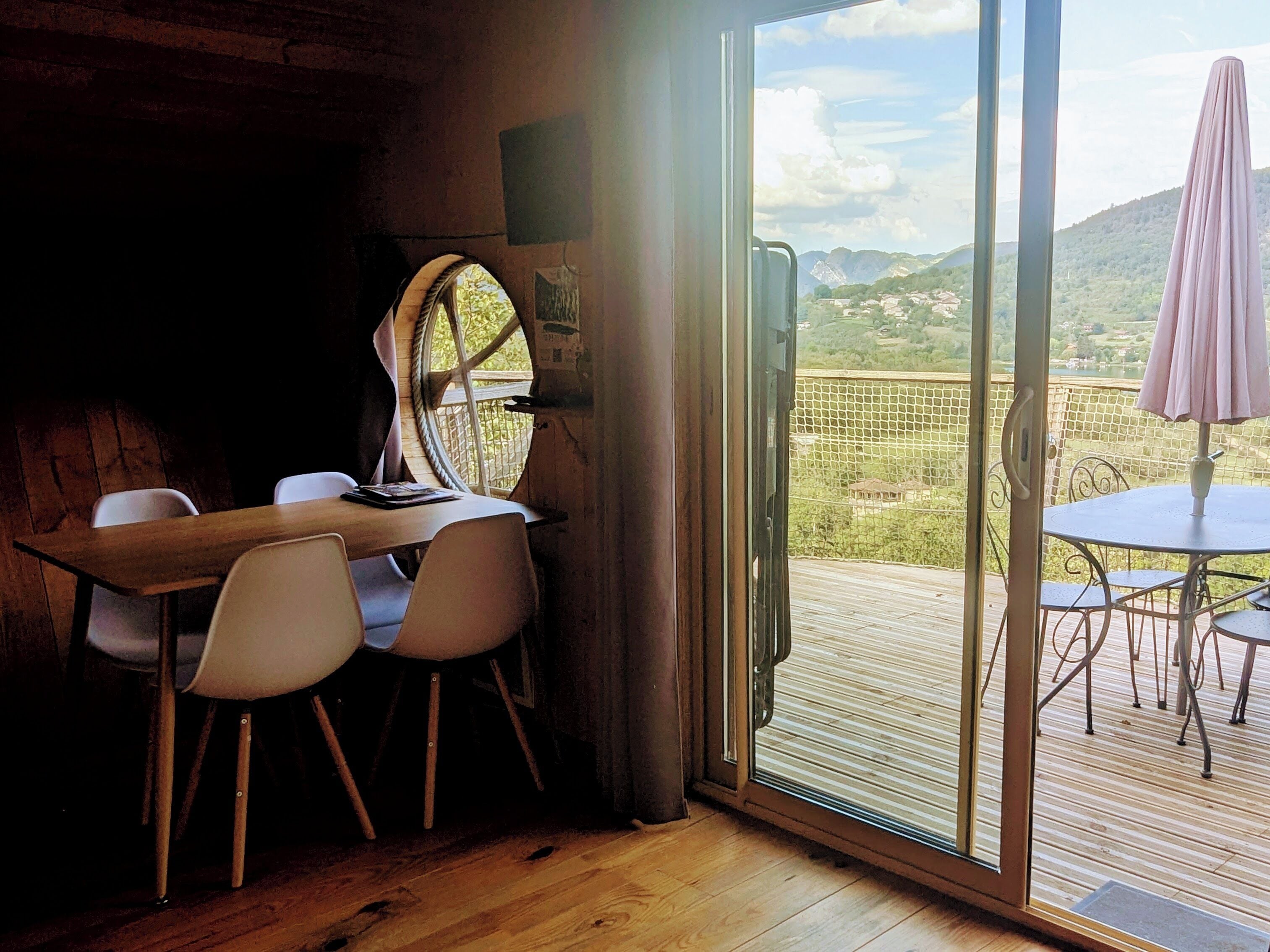 A wooden interior with a table and chairs, a large window with a view of a green landscape, and a balcony with a table and chairs.