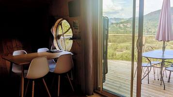 A wooden interior with a table and chairs, a large window with a view of a green landscape, and a balcony with a table and chairs.