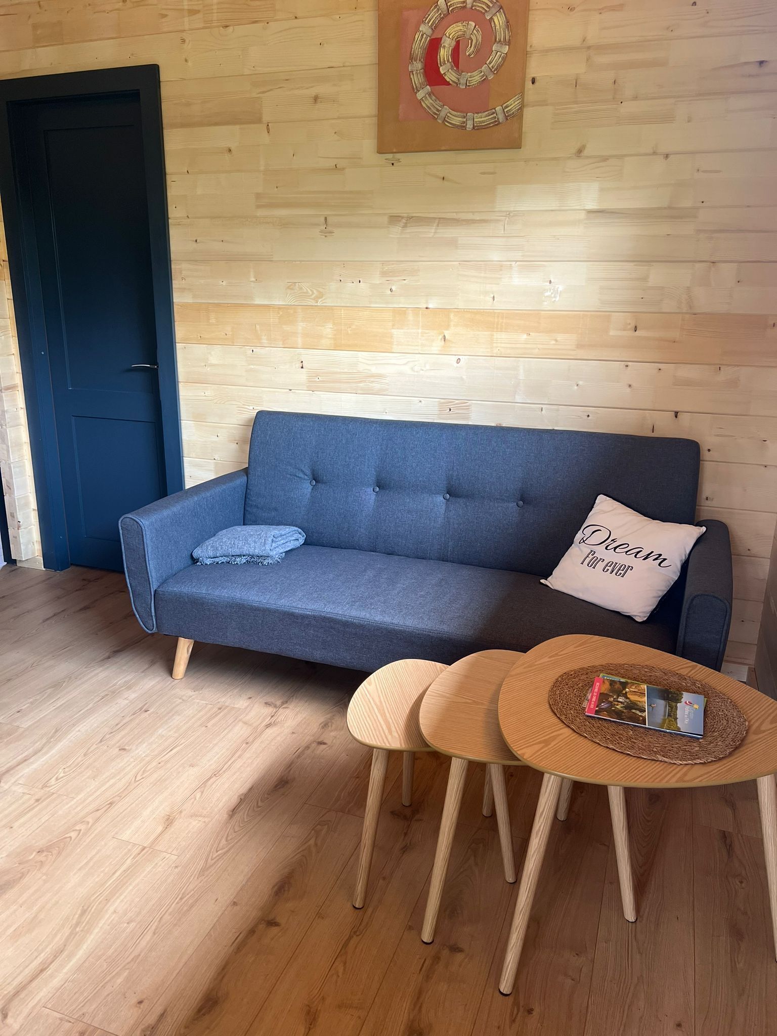 Una habitación con un sofá azul, una pequeña mesa de madera con libros, y una pared de madera con un diseño circular.