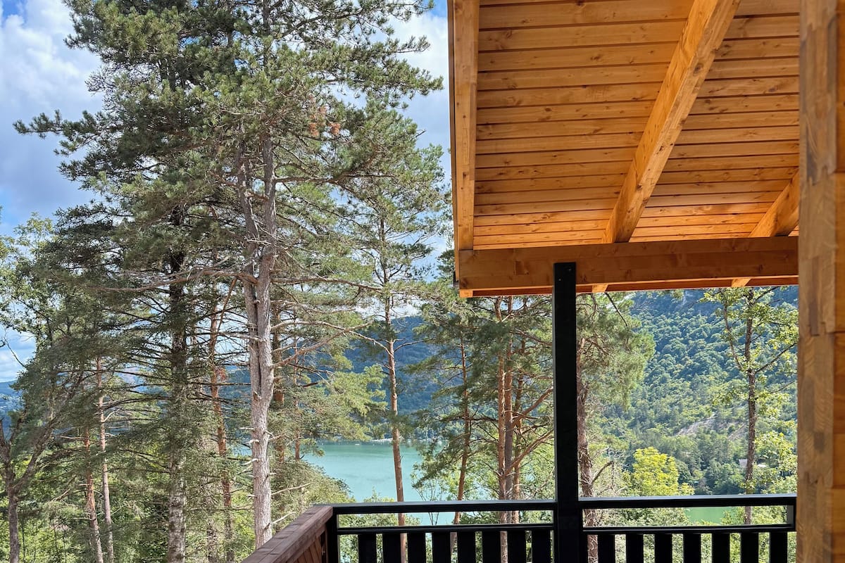 Une terrasse en bois dotée d’une rambarde, offrant une vue sur une zone boisée et un plan d’eau.