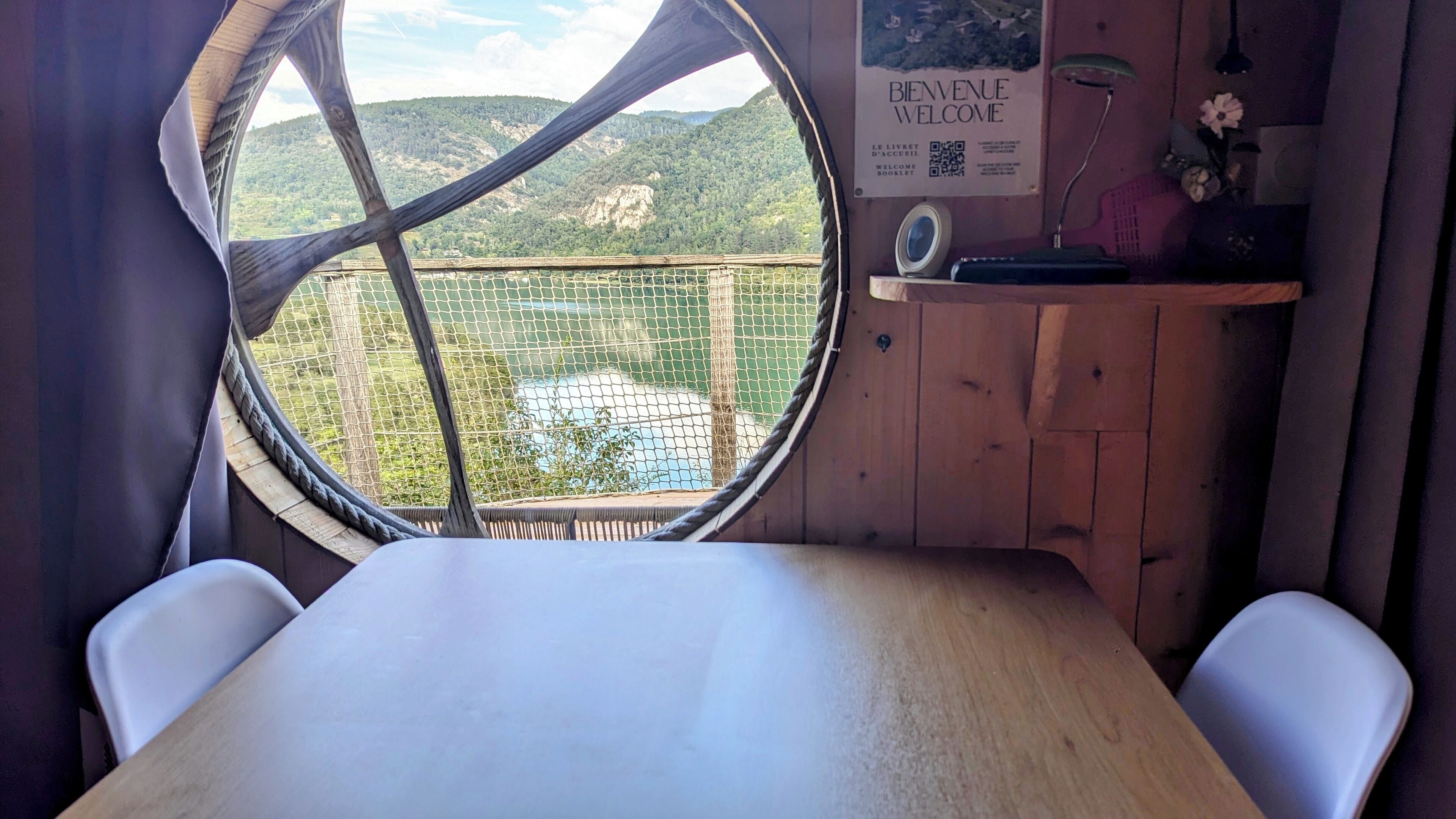 A room with a wooden table, two white chairs, a round window with a view of a green landscape and a mountain.