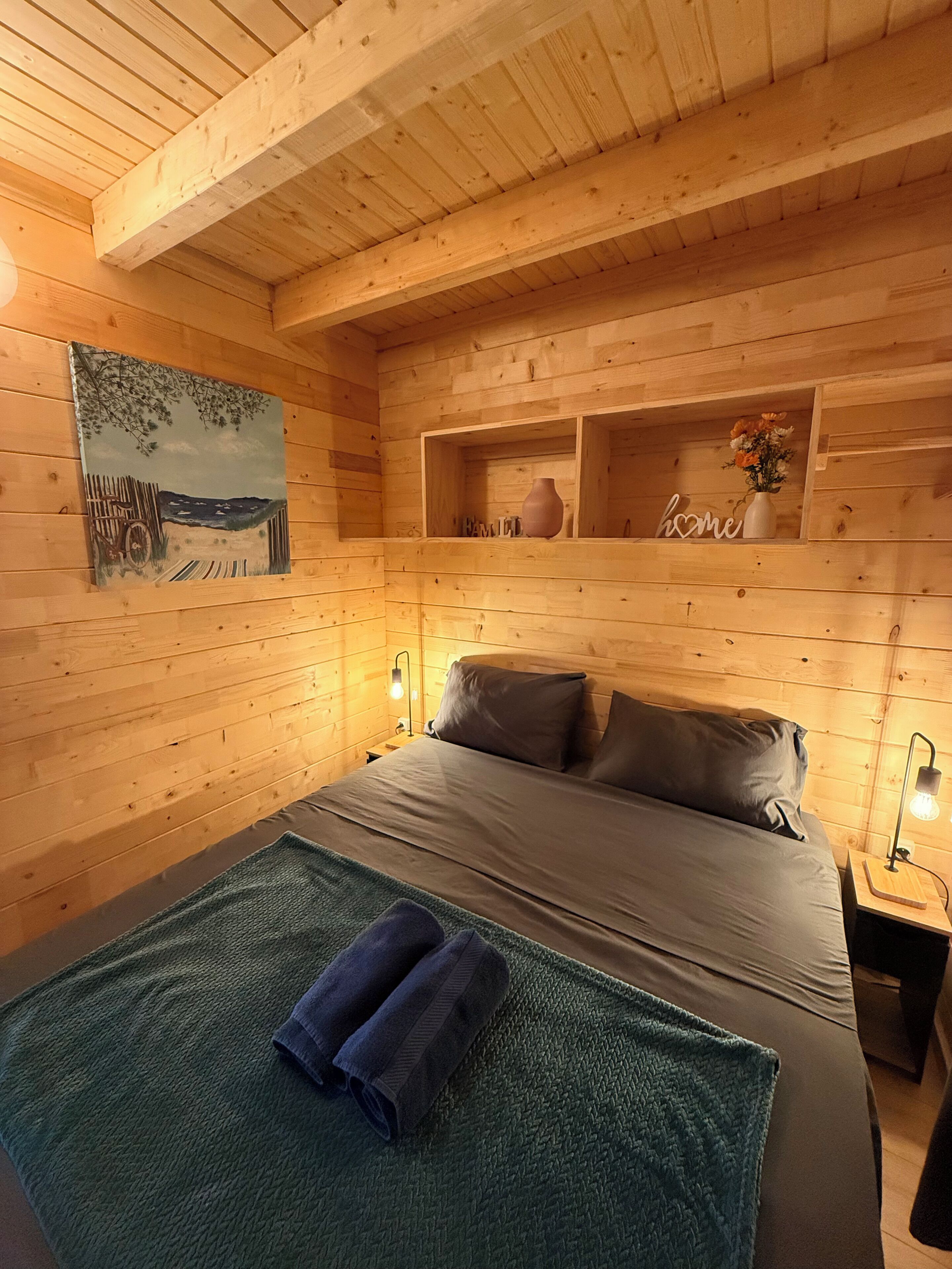 Un dormitorio de cabaña de madera con una cama, almohadas, un cuadro y estantes con adornos.