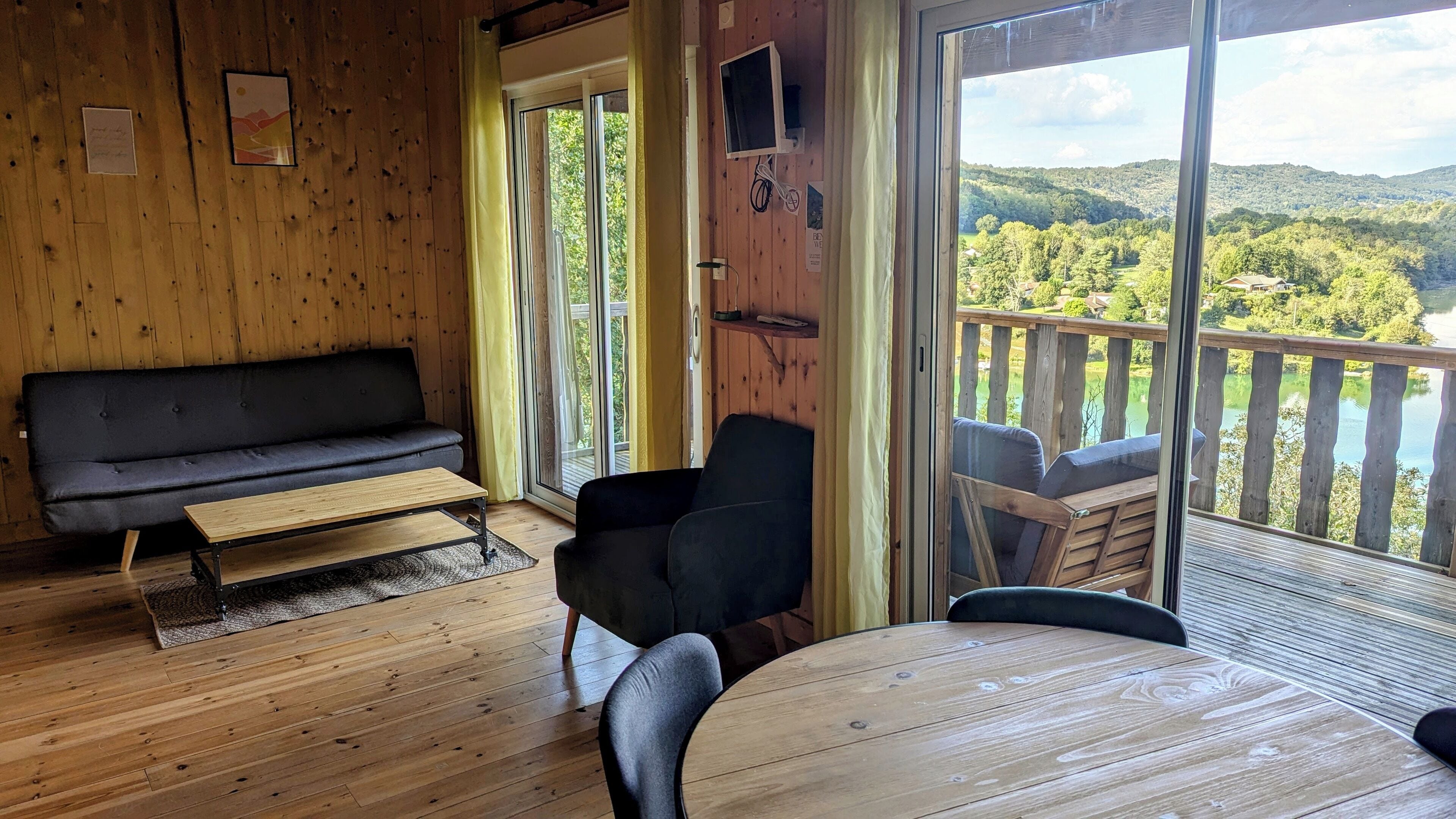 A wooden cabin interior with a sofa, a table, and chairs. There is a large window with a view of a lake and hills.