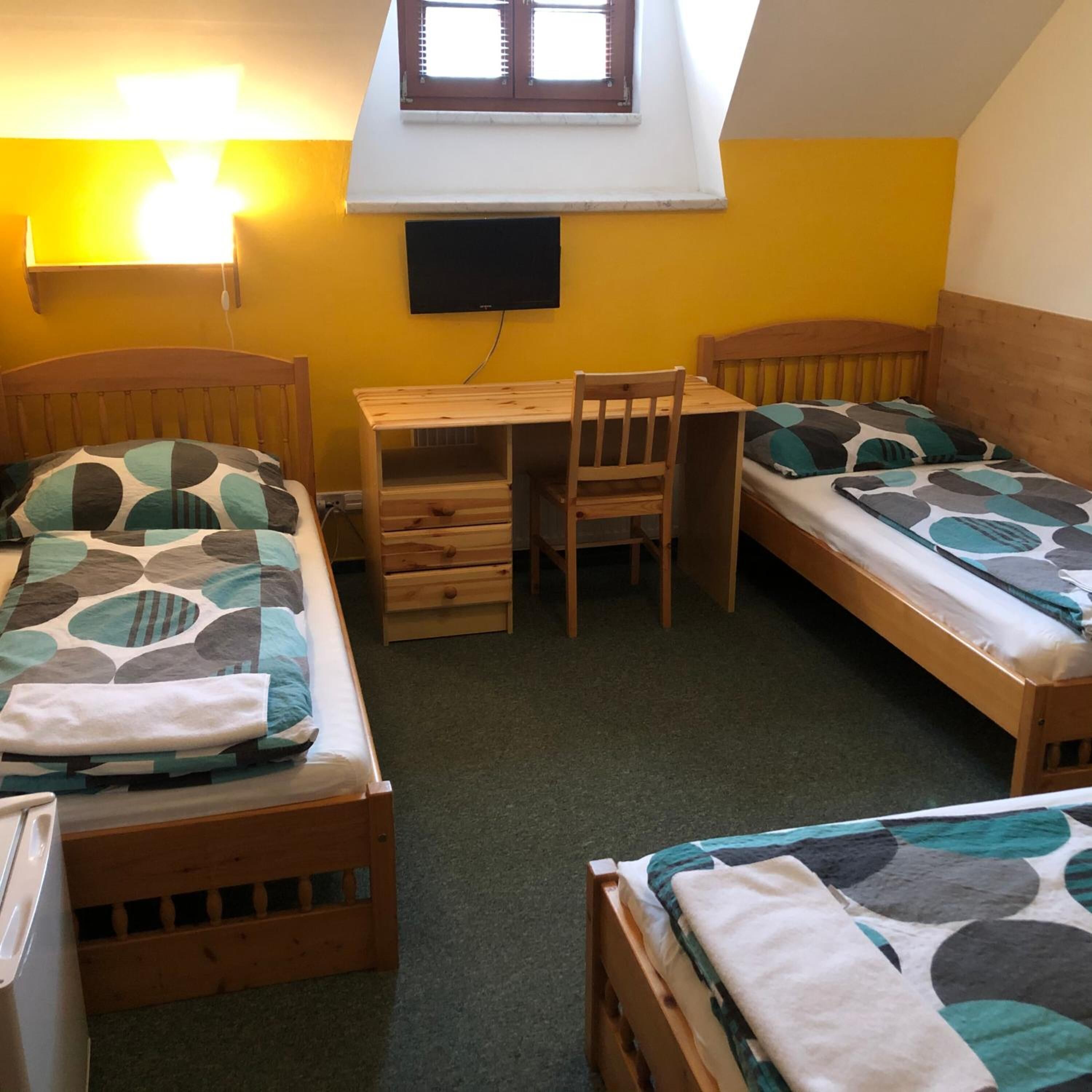 A dormitory room with two single beds, a wooden desk, a chair, and a TV mounted on the wall.