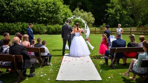 Una ceremonia de boda al aire libre con invitados sentados en bancos, un pasillo blanco y follaje en el fondo.