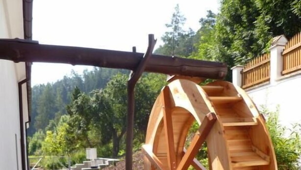Una estructura de rueda hidráulica de madera con una cuenca rectangular, ubicada en un jardín con un camino pavimentado y follaje en el fondo.