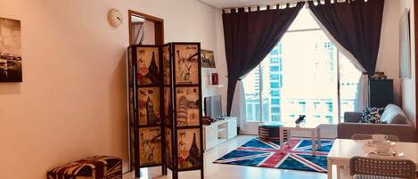 A modern living room with a large window, a Union Jack rug, a sofa, a dining table with chairs, and a decorative screen with travel-themed artwork.