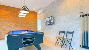 Uma mesa de bilhar com tecido azul, duas cadeiras dobráveis, uma televisão fixada na parede e um suporte com tacos de bilhar.