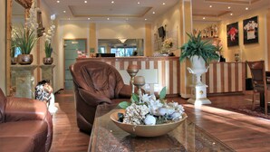 A lobby with a reception desk, leather chairs, a large decorative bowl with flowers, and a mirror with an ornate frame. - Apart Hotel Scheuer (Huerth)