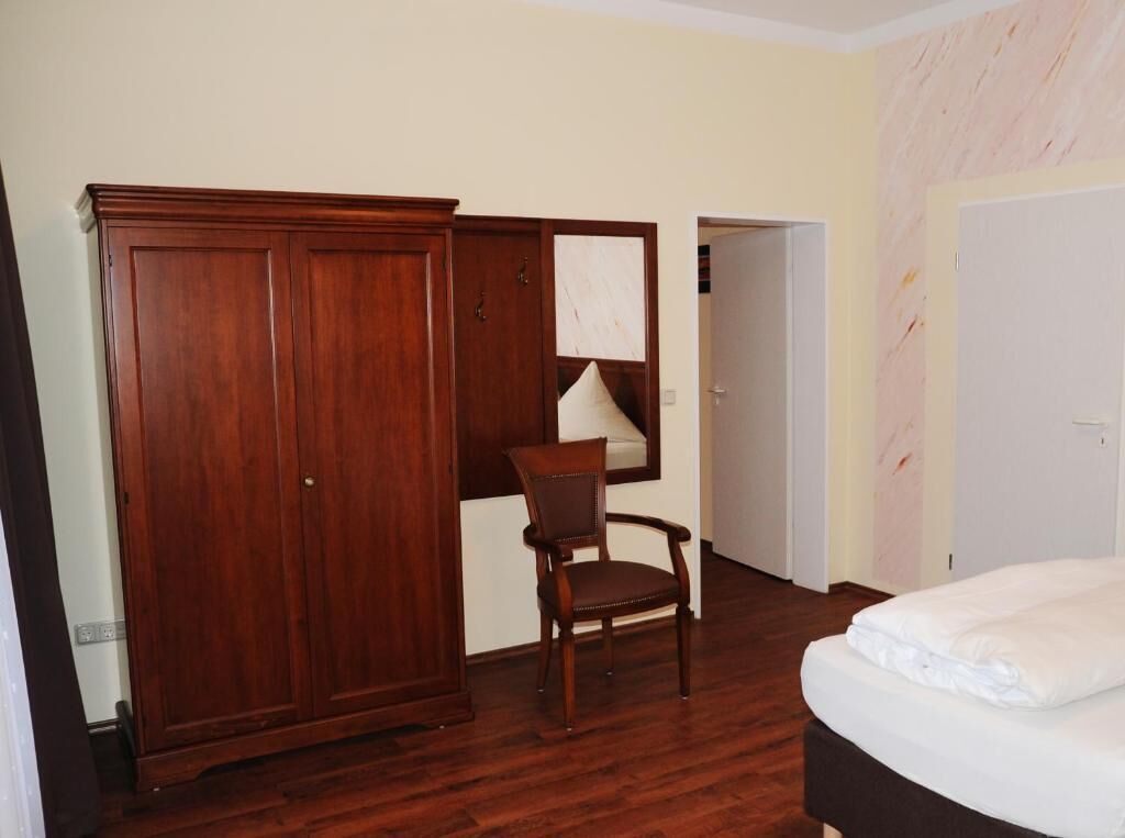 A bedroom with a wooden wardrobe, a chair, a bed, and a door.