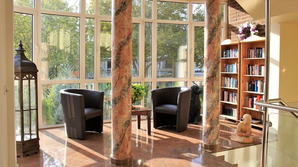 A spacious lobby with large windows, marble columns, a fireplace, and a bookshelf. - Apart Hotel Scheuer (Huerth)