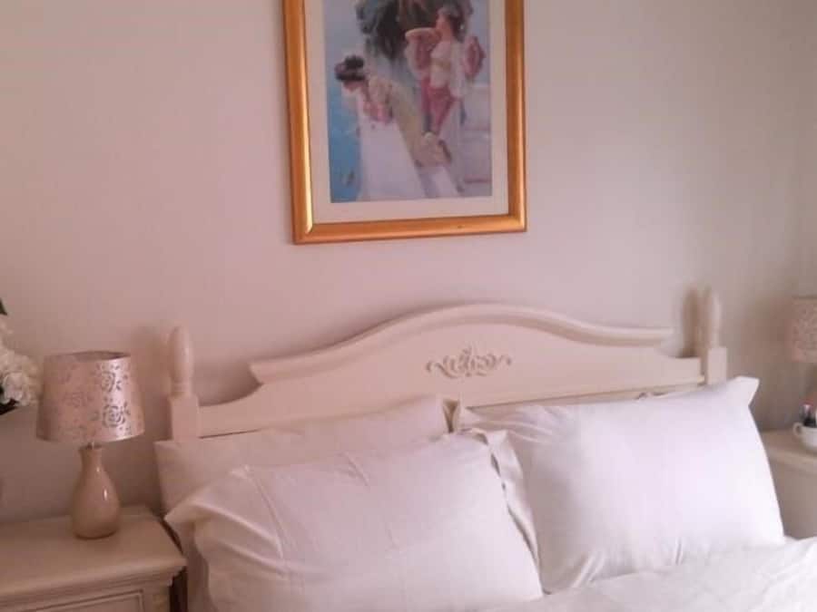 Un dormitorio con cama blanca, mesita de noche con lámpara y un cuadro en la pared.