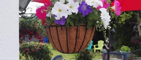 Una maceta colgante con flores blancas, moradas y rosas, un paraguas rojo y mobiliario de exterior.