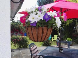A hanging flower basket with white, purple, and pink flowers, a red umbrella, and outdoor furniture.