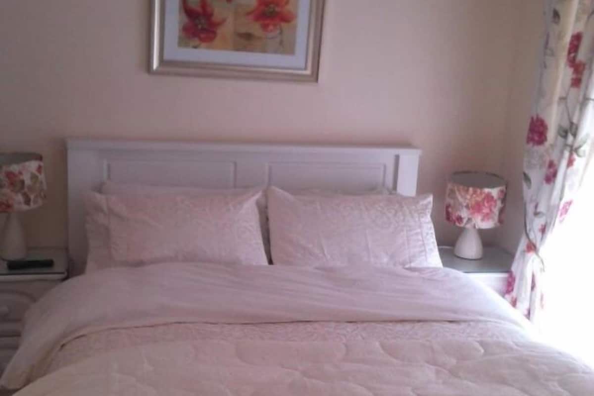 A bed with a white headboard, a floral painting on the wall, and a nightstand with a lamp.