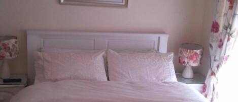 A bed with a white headboard, a floral painting on the wall, and a nightstand with a lamp.