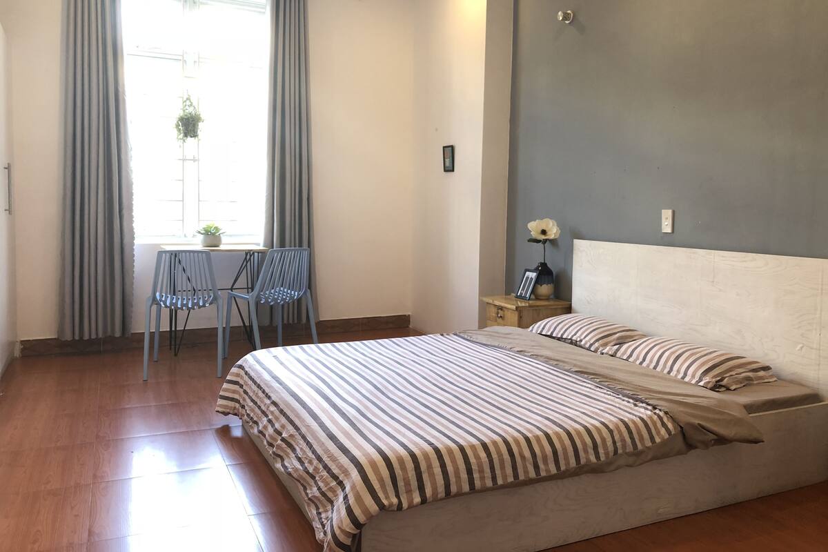 A bedroom with a bed, a chair, a nightstand, a window with curtains, and a potted plant.