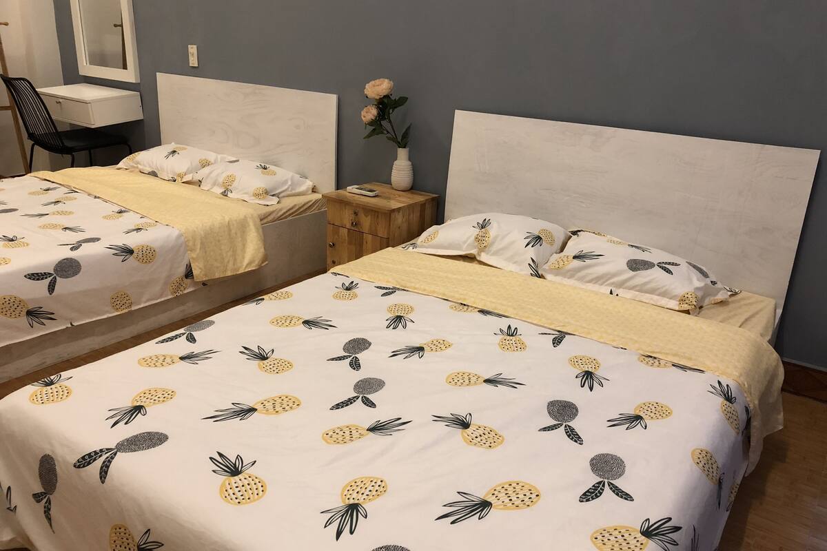 A bedroom with two beds, each with a pineapple-themed bedspread, a wooden nightstand, a white vase with flowers, and a mirror on the wall.